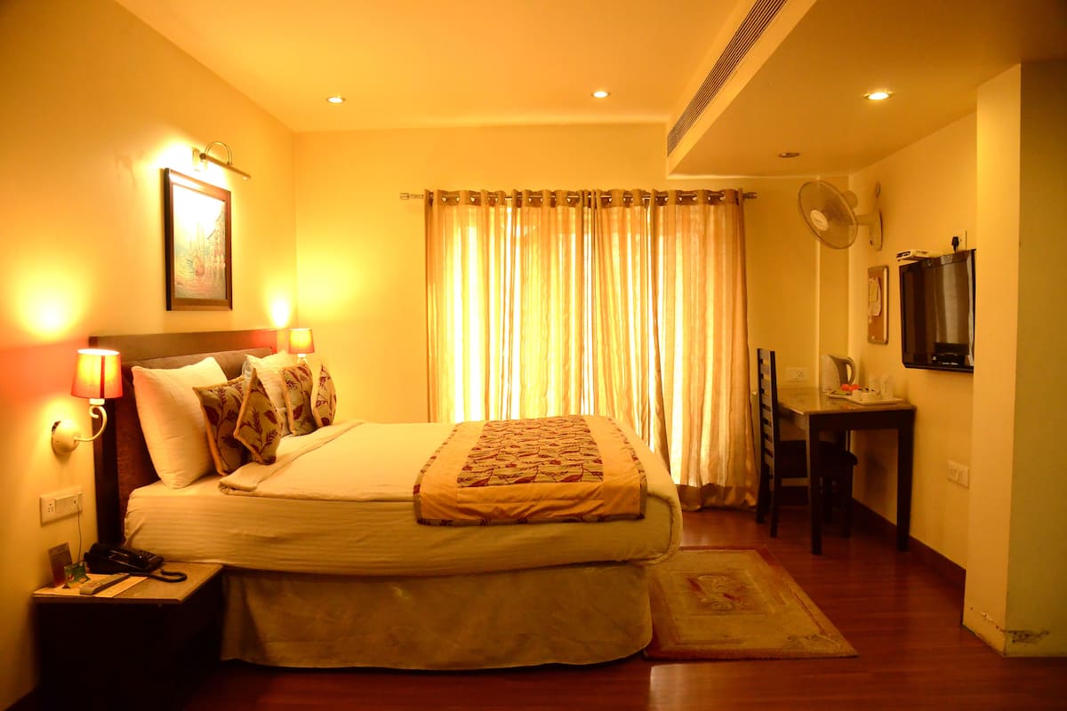 A hotel room with a bed, bedside lamps, a desk, a chair, a fan, and a television.