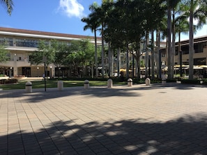 A spacious courtyard with a tiled floor, surrounded by palm trees and a building with a red-tiled roof. - LYX Suites at Merrick Park in Coral Gables (Coral Gables)
