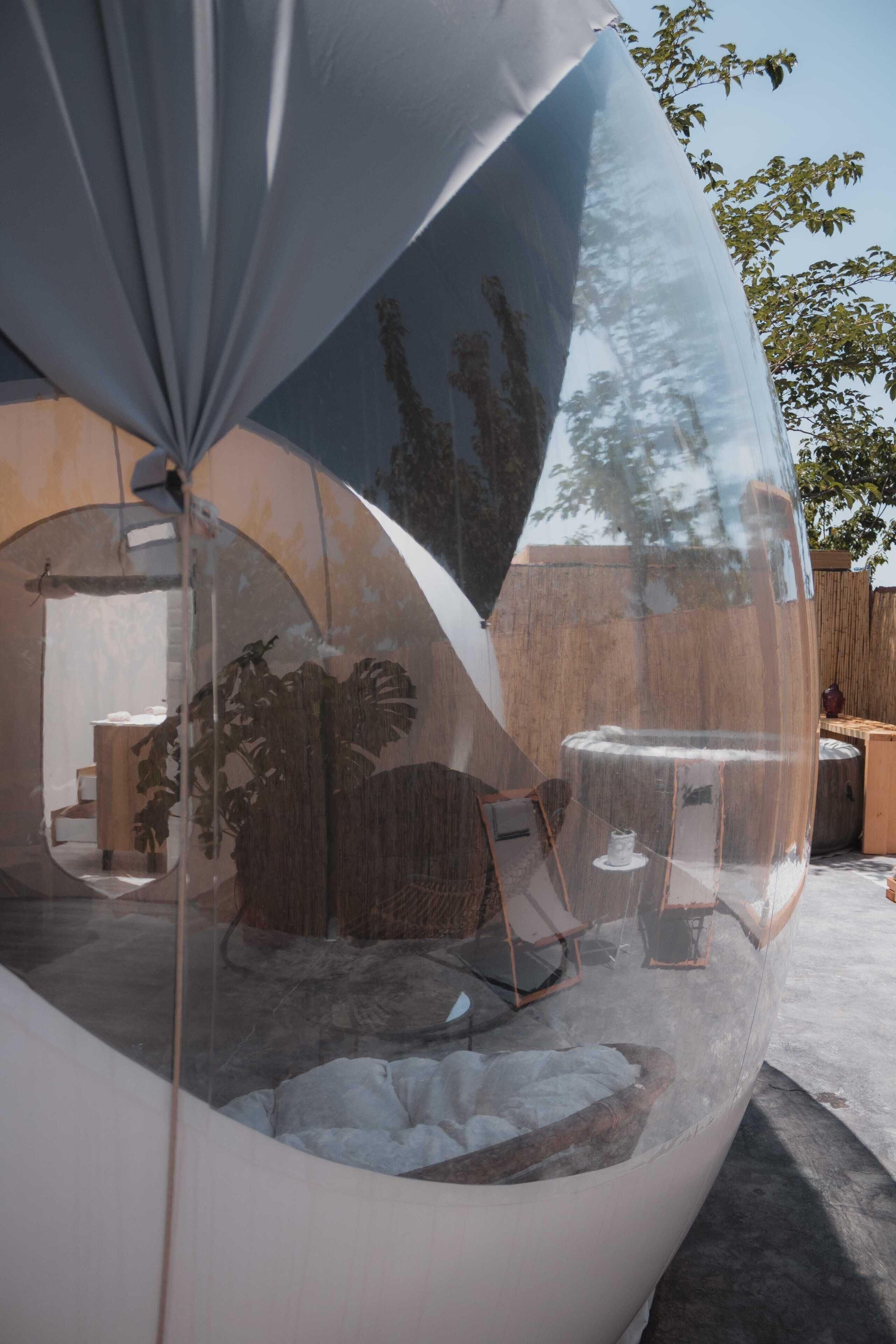 Una estructura de cúpula transparente con un interior visible que incluye una mesa y sillas, rodeada por una base de hormigón y una valla de madera al fondo.