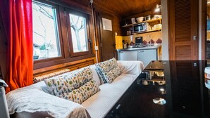 A cozy cabin interior with a sofa, a dining table, a kitchen area, and large windows with curtains. - Fuente del Lobo Glamping  Bungalows - Adults Only (Pinos Genil)