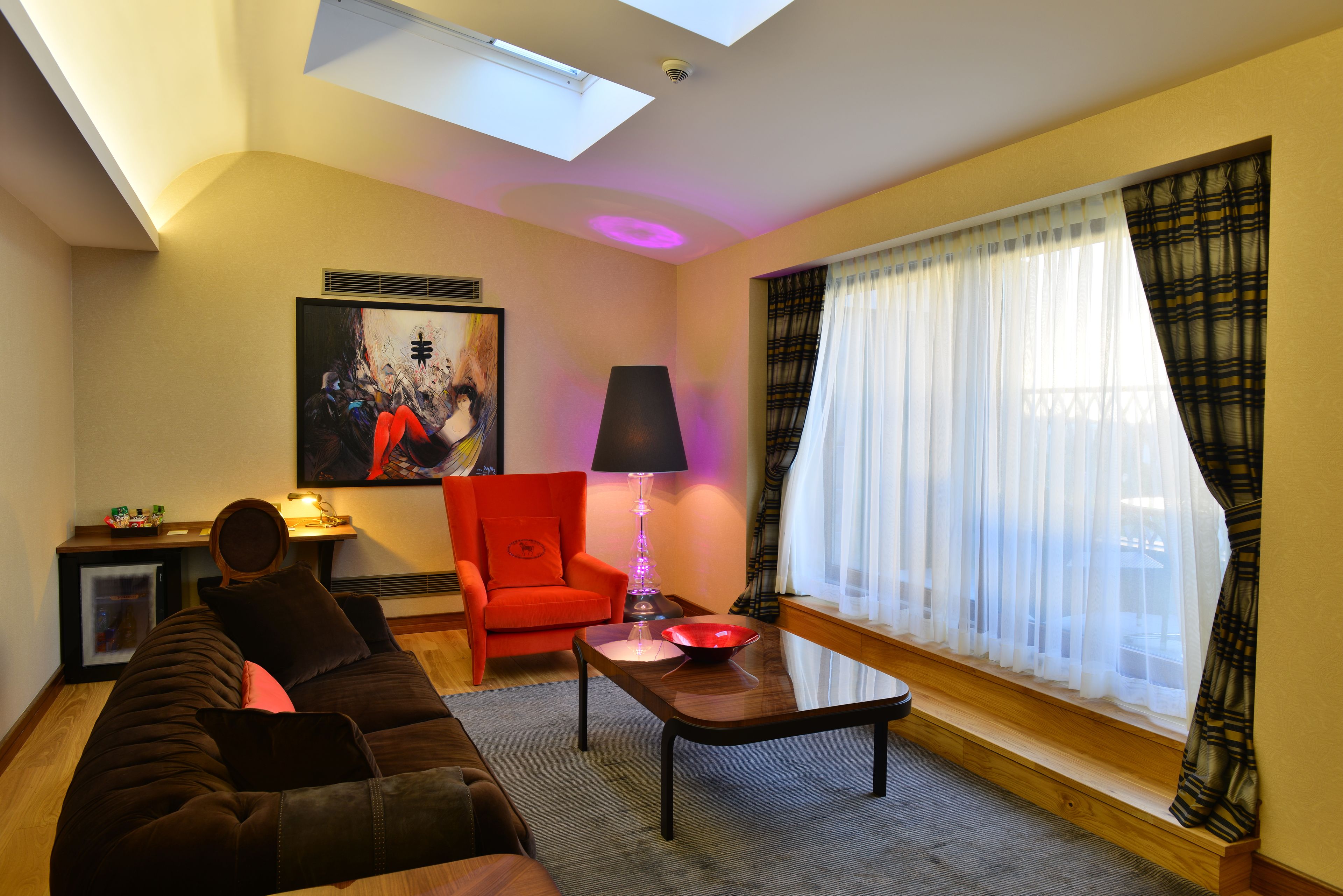 A living room with a sofa, a red armchair, a table with a lamp, a painting on the wall, and a window with sheer curtains.