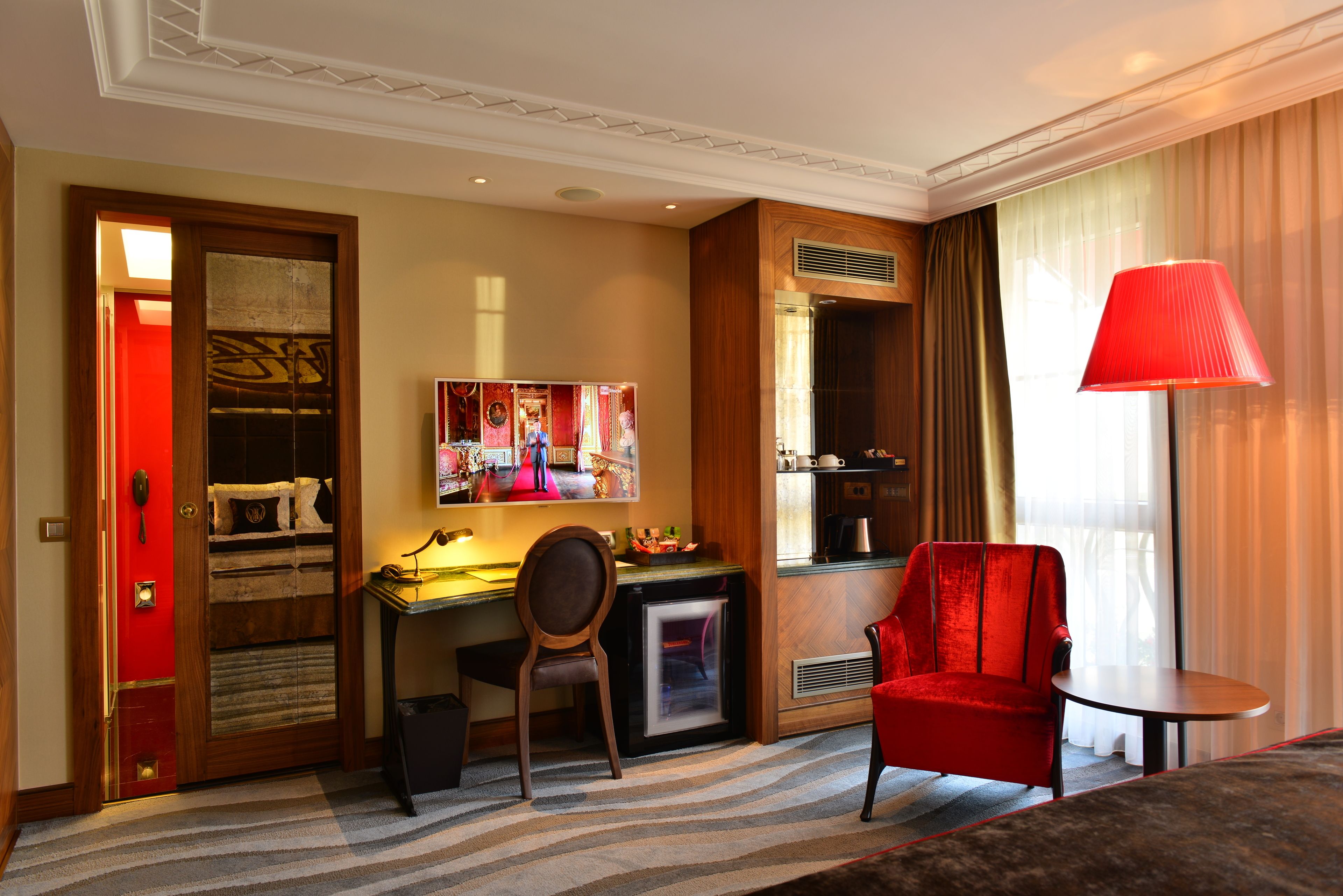 Habitación de hotel con escritorio, una silla roja, una mesita, una lámpara roja y un ventanal grande con cortinas.
