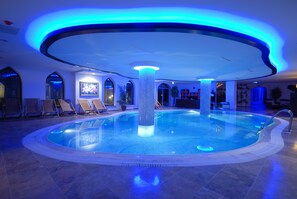 Une grande piscine intérieure dotée d’un pilier central, entourée de chaises longues et agrémentée d’un design de plafond moderne.