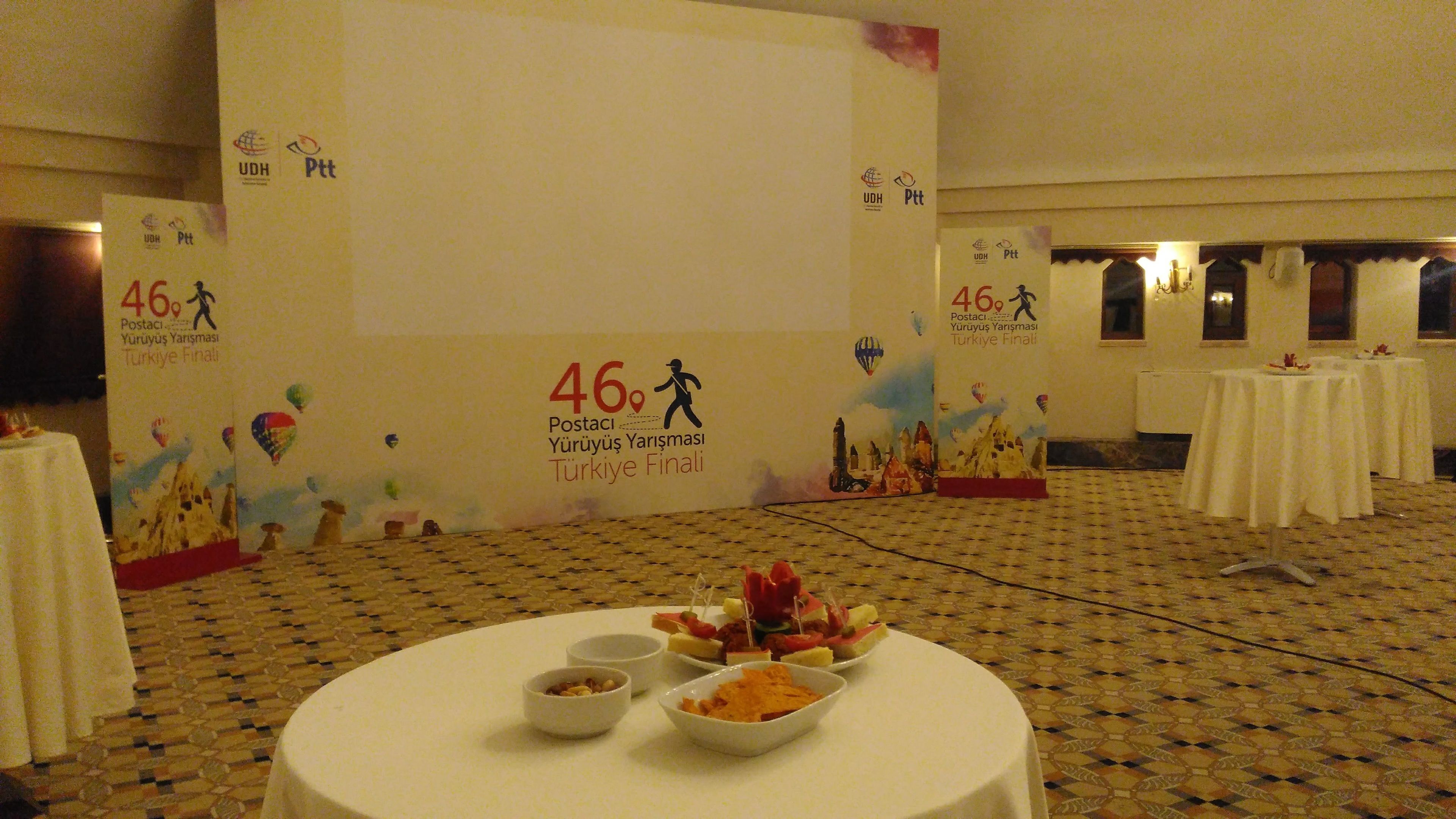 A conference room with a large screen displaying '46 Postaci Yürüyüş Yarışması Türkiye Finali' and tables with food.