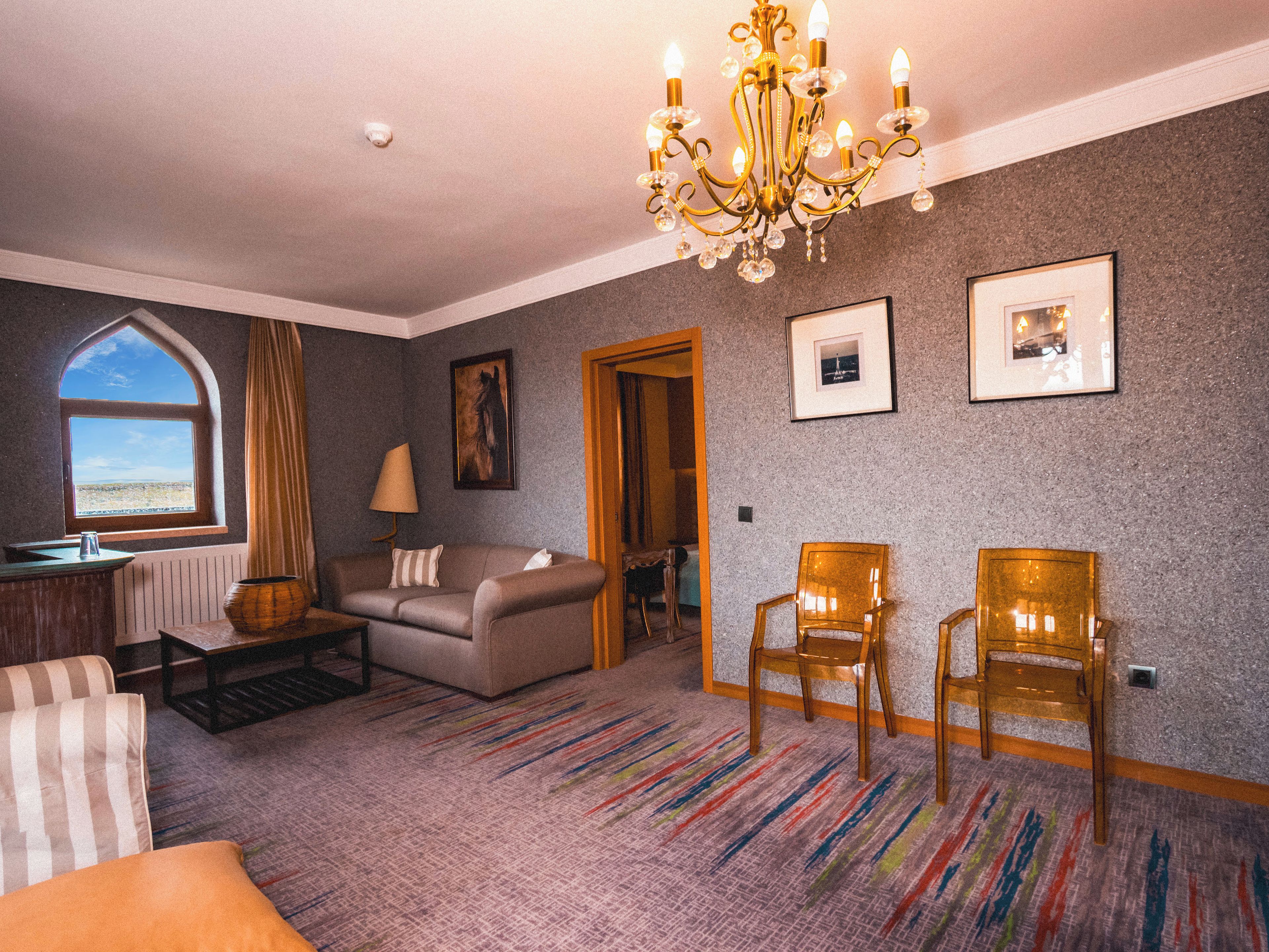A room with a large window, a chandelier, a sofa, two chairs, and a carpeted floor.