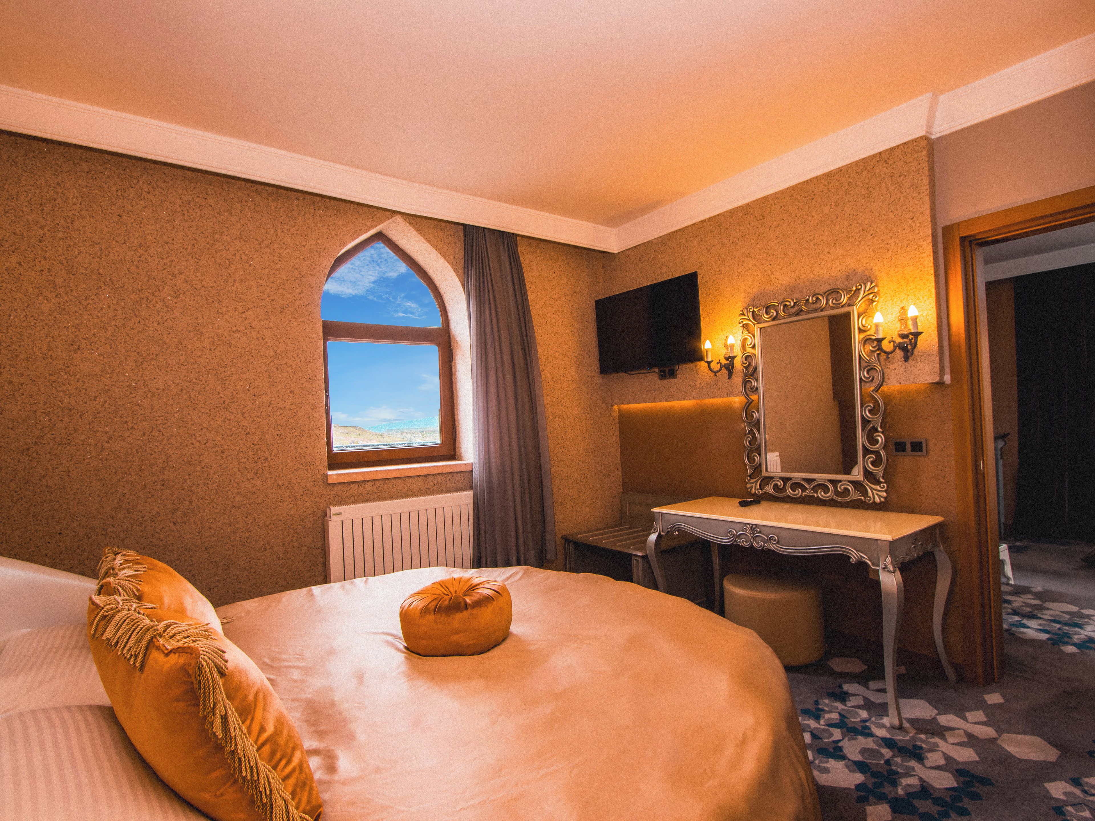 Een hotelkamer met een groot bed, een kaptafel met spiegel, een televisie aan de muur en een raam met uitzicht op de lucht.