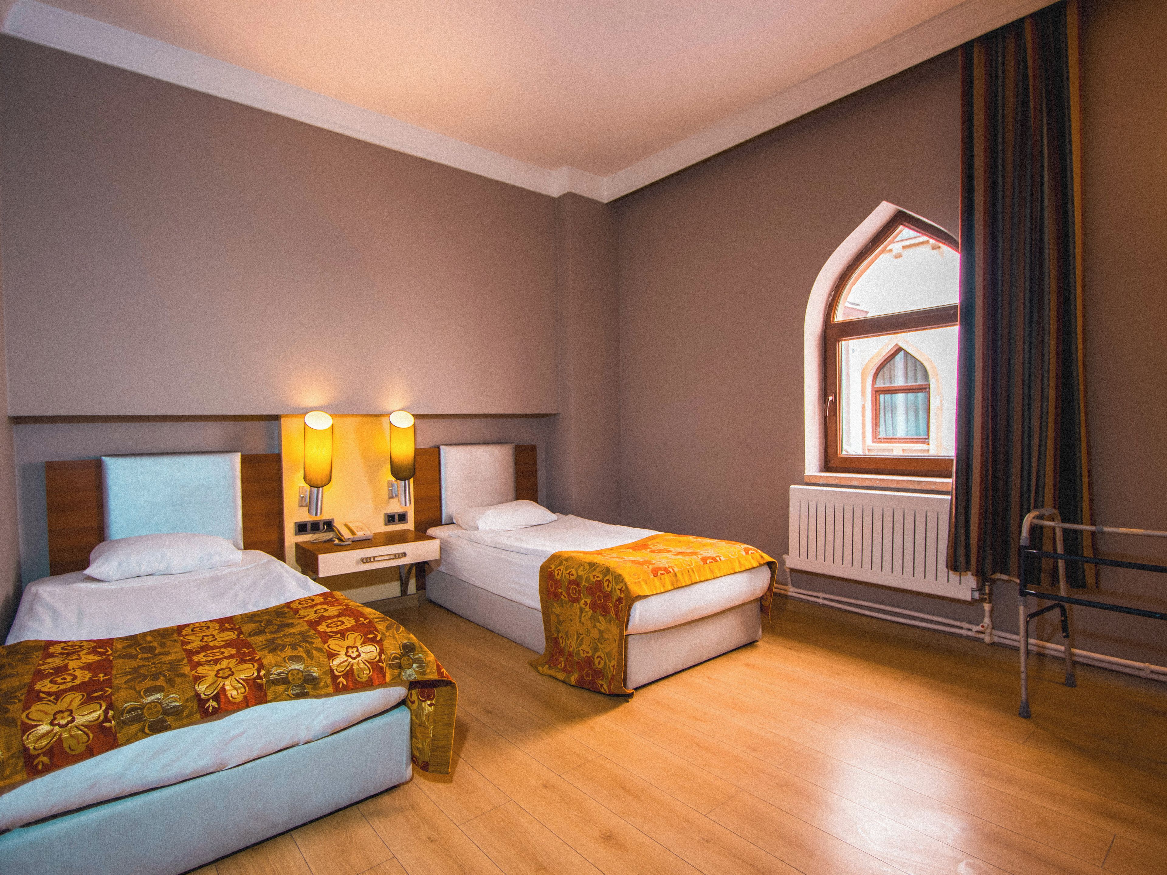 A hotel room with two beds, a nightstand with lamps, a window with a view, and a radiator.