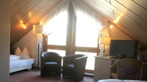 A cozy attic room with a bed, two armchairs, a desk with a TV, and a large window with curtains.