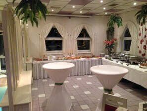 A banquet hall with a long table set for a meal, two round tables with white coverings, and a window with a view of the night sky.