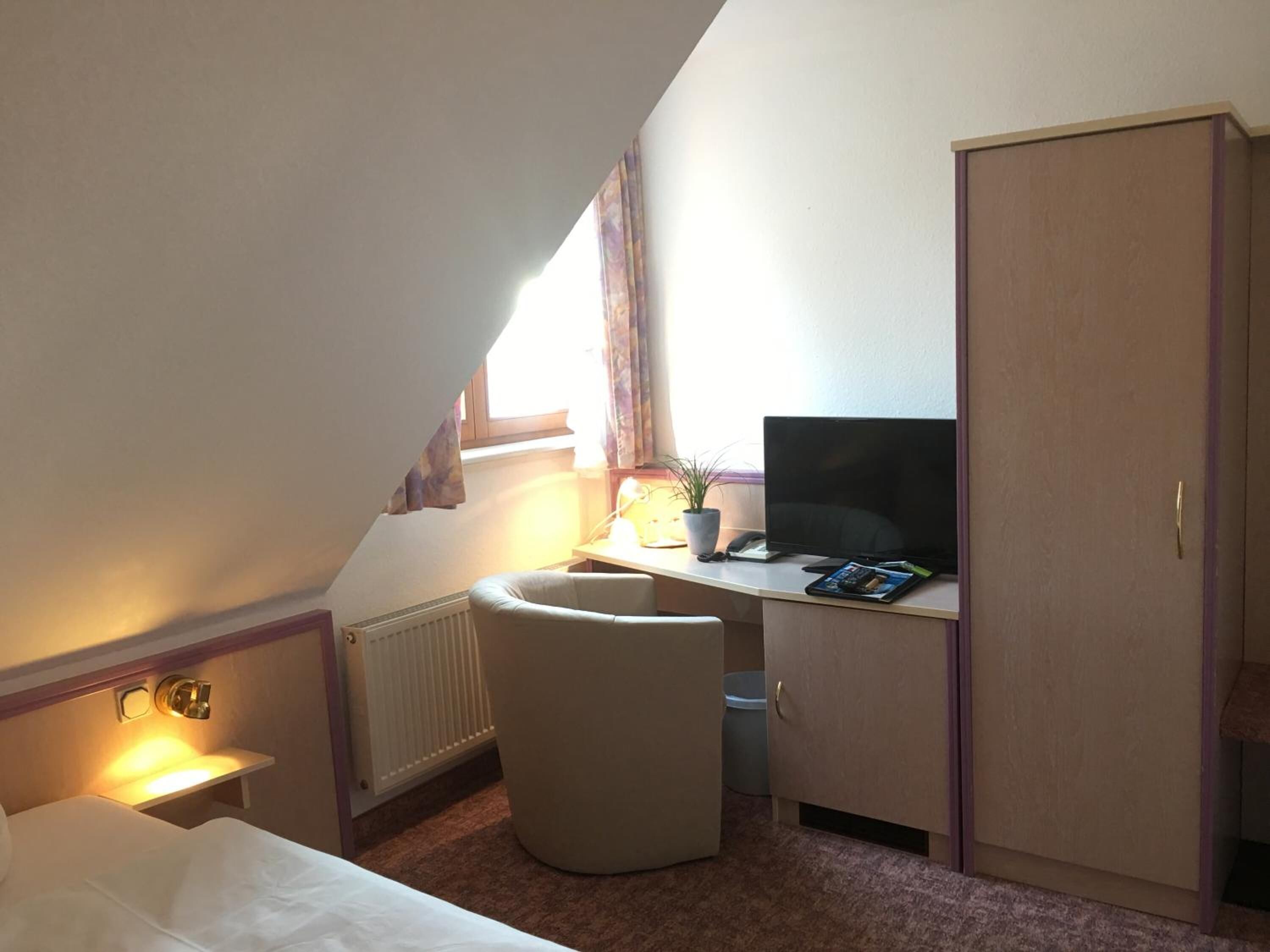 A small, well-lit room with a bed, a desk with a computer, a chair, and a wardrobe.