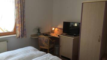 Een kamer met twee bedden, een bureau met computer en televisie, een stoel, een potplant en een garderobe.