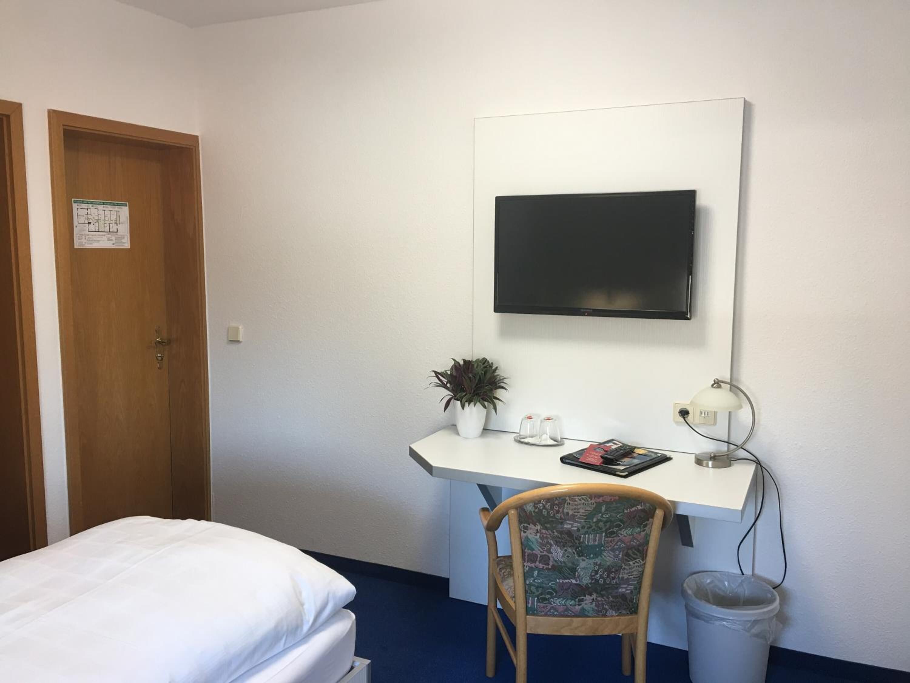 A hotel room with a bed, a desk with a computer, a chair, a TV mounted on the wall, and a trash can.