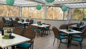 A covered outdoor dining area with wicker chairs and tables, potted plants, and green paper lanterns.