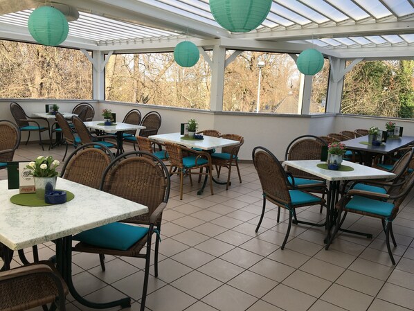 A covered outdoor dining area with wicker chairs and tables, potted plants, and green paper lanterns.