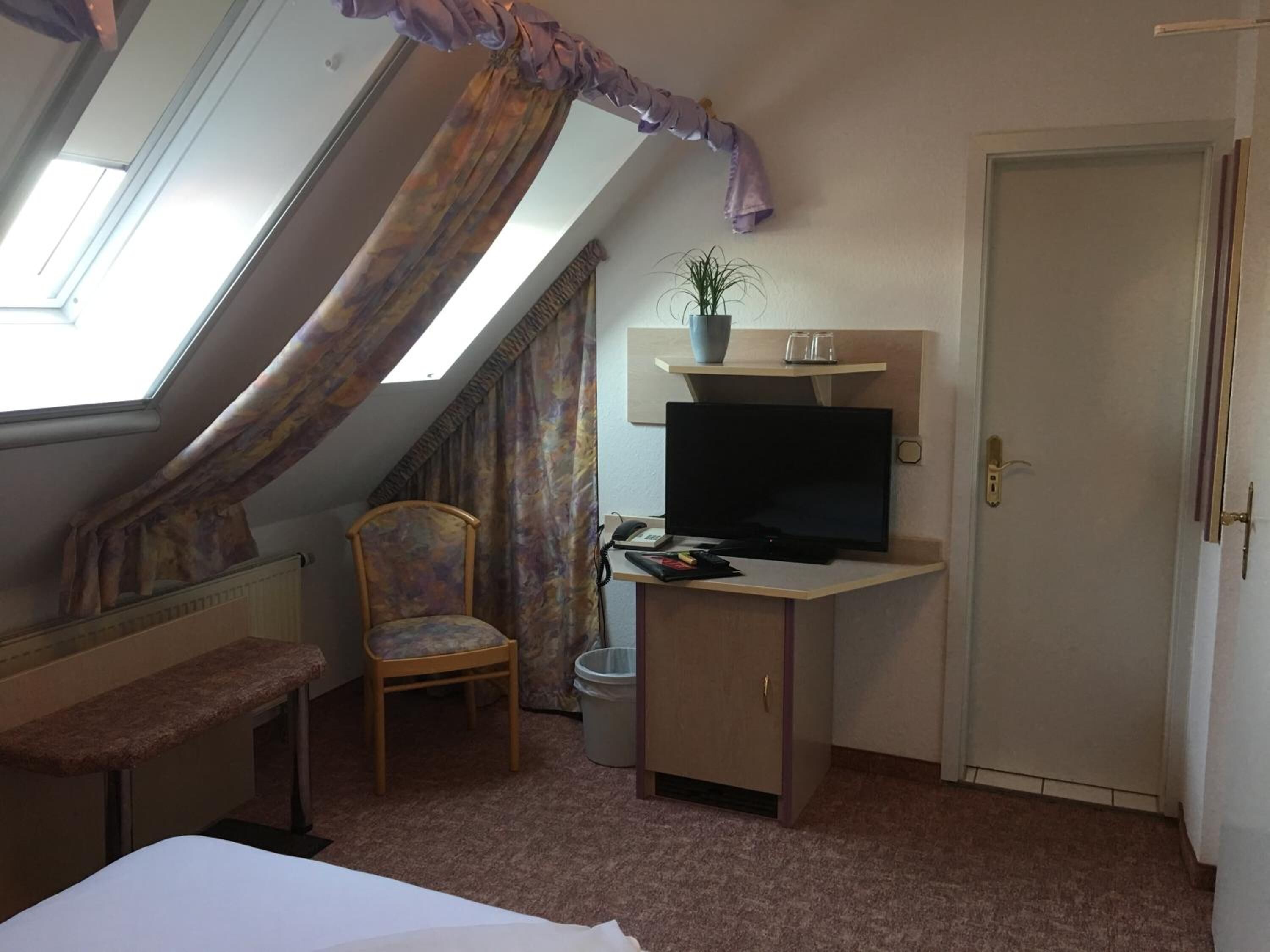 Ein kleiner Raum mit schrägem Dach, einem Bett, einem Sessel, einem Schreibtisch mit Fernseher, einer Zimmerpflanze und einer Tür.