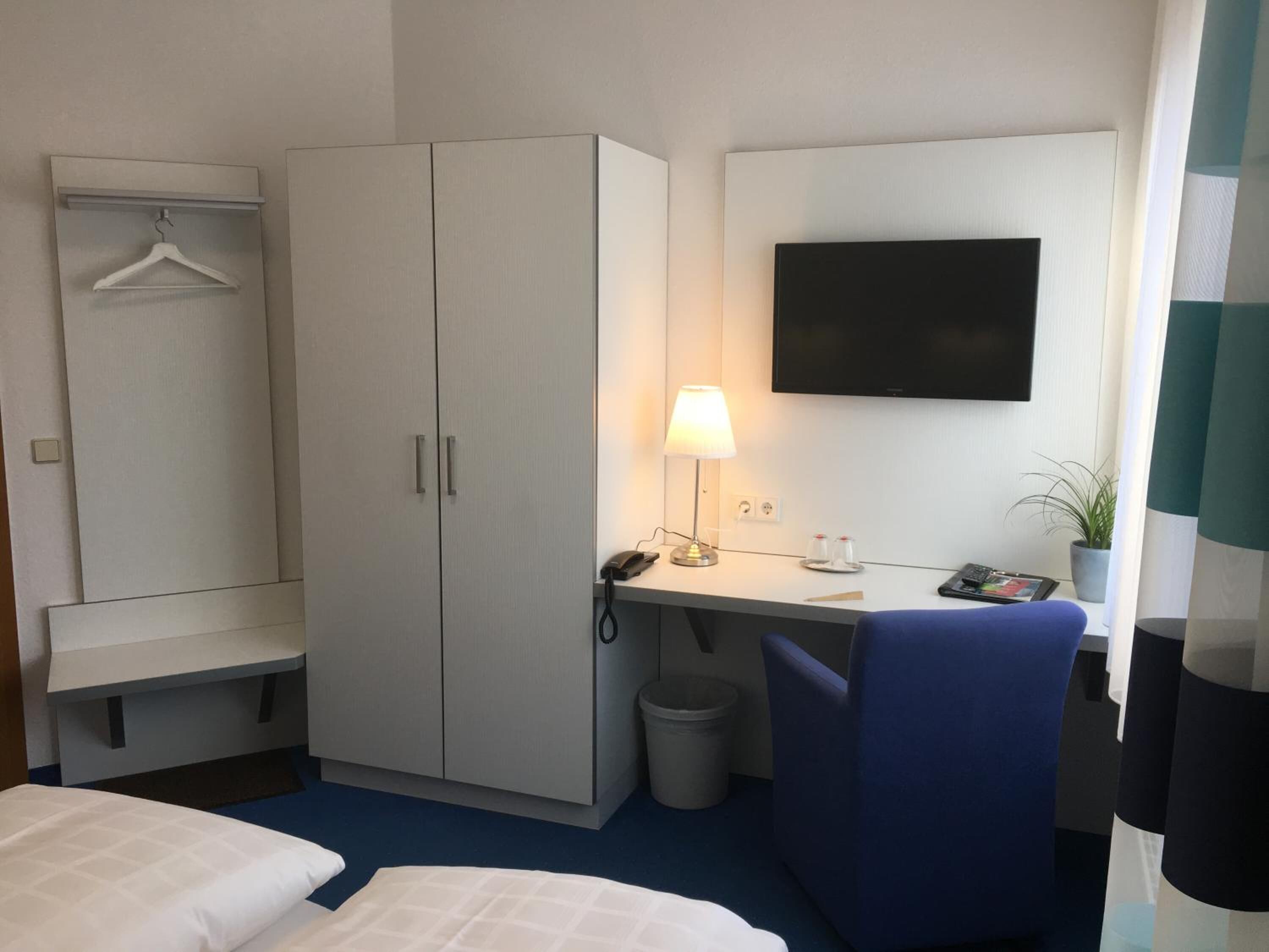 Habitación de hotel con cama, escritorio con lámpara, televisor, armario y cubo de basura.