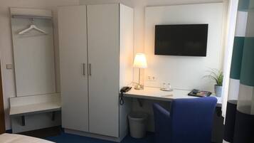 Habitación de hotel con cama, escritorio con lámpara, televisor, armario y cubo de basura.