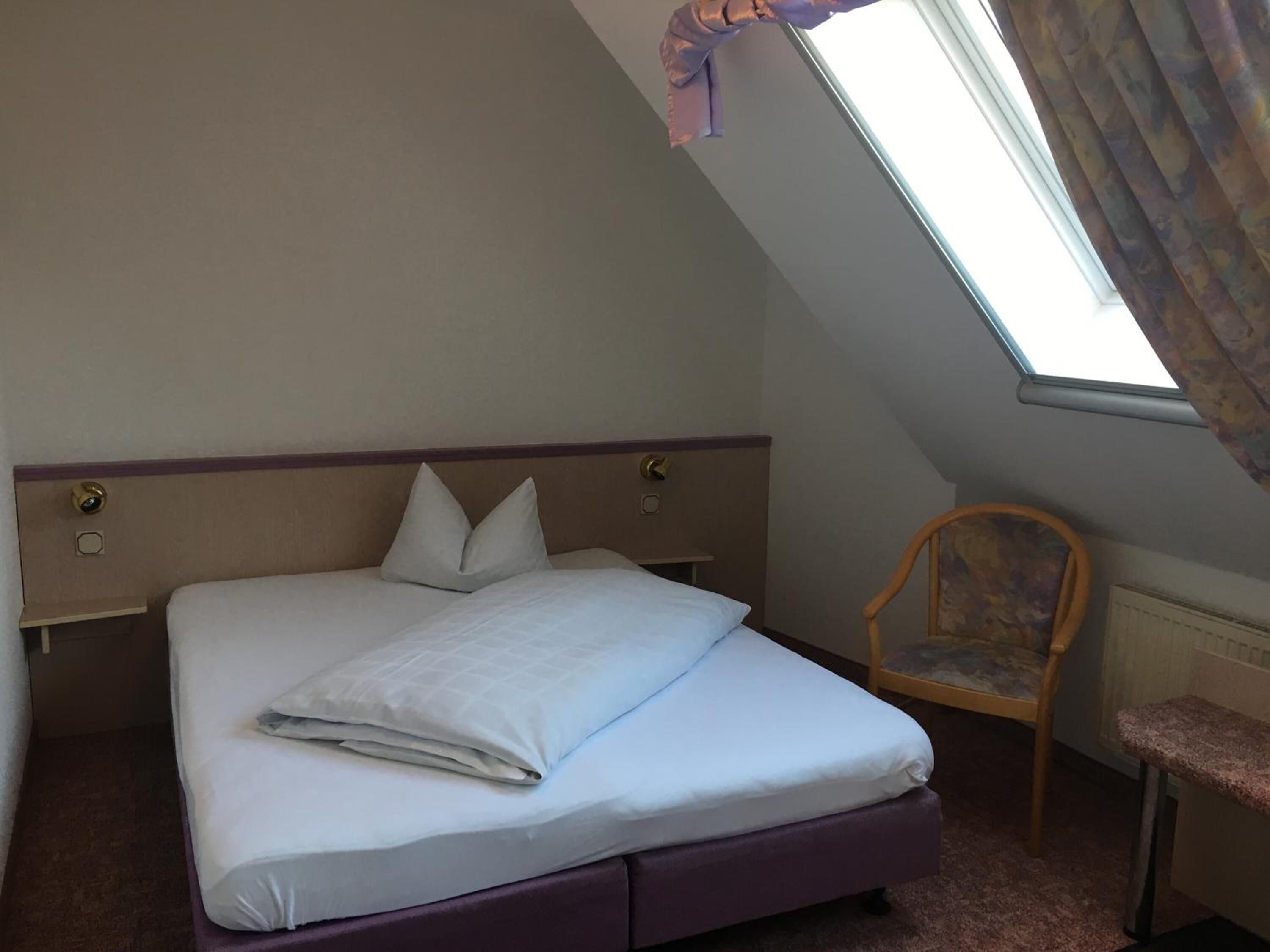 Una cama bien tendida con almohadas, una silla de madera y una mesita en una habitación con techo inclinado y una ventana.