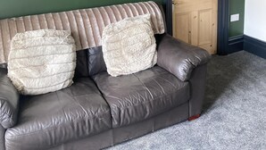 A dark brown leather sofa with two fluffy pillows against a green wall, a wooden door, and a carpeted floor. - Brinkburn Serviced Apartments (Darlington)