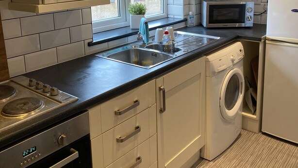 A modern kitchen with white cabinets, a built-in oven, a microwave, and a washing machine. - Brinkburn Serviced Apartments (Darlington)