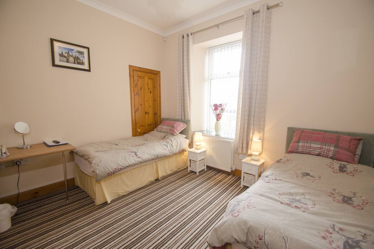 A bedroom with two beds, a desk, a window with curtains, and a radiator.