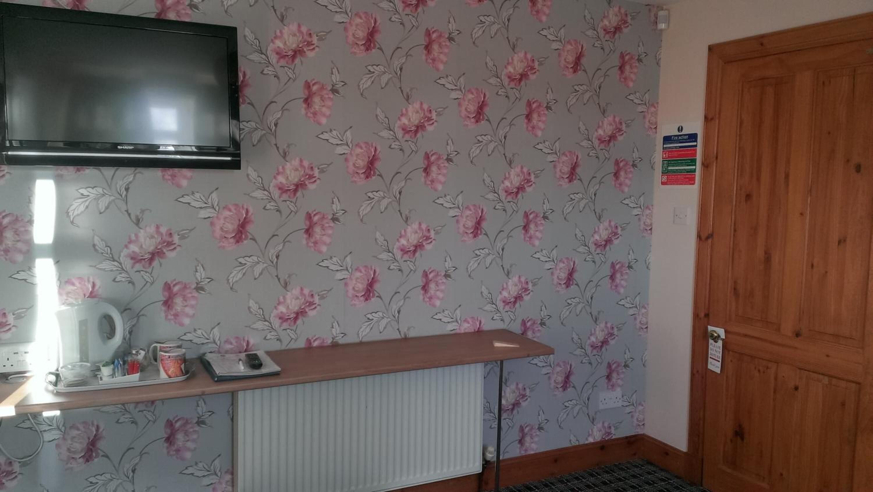 A room with a floral wallpaper, a wooden door, a radiator, a TV mounted on the wall, and a small table with a mug and remote control.