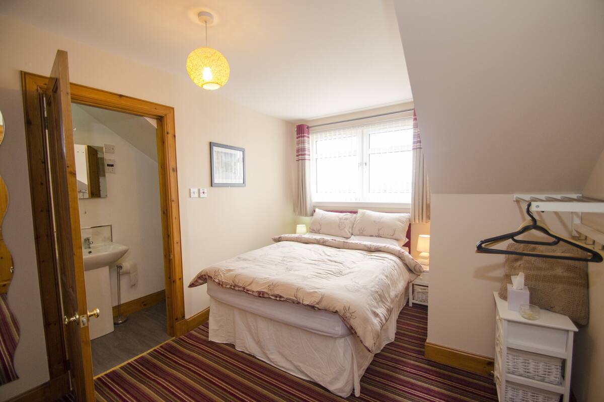 A bedroom with a bed, a bedside table, a hanging light fixture, a window with curtains, and a door leading to a bathroom.