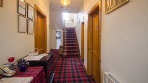 A hallway with a red and black plaid carpet, wooden doors, framed certificates on the wall, and a staircase leading to an upper floor.