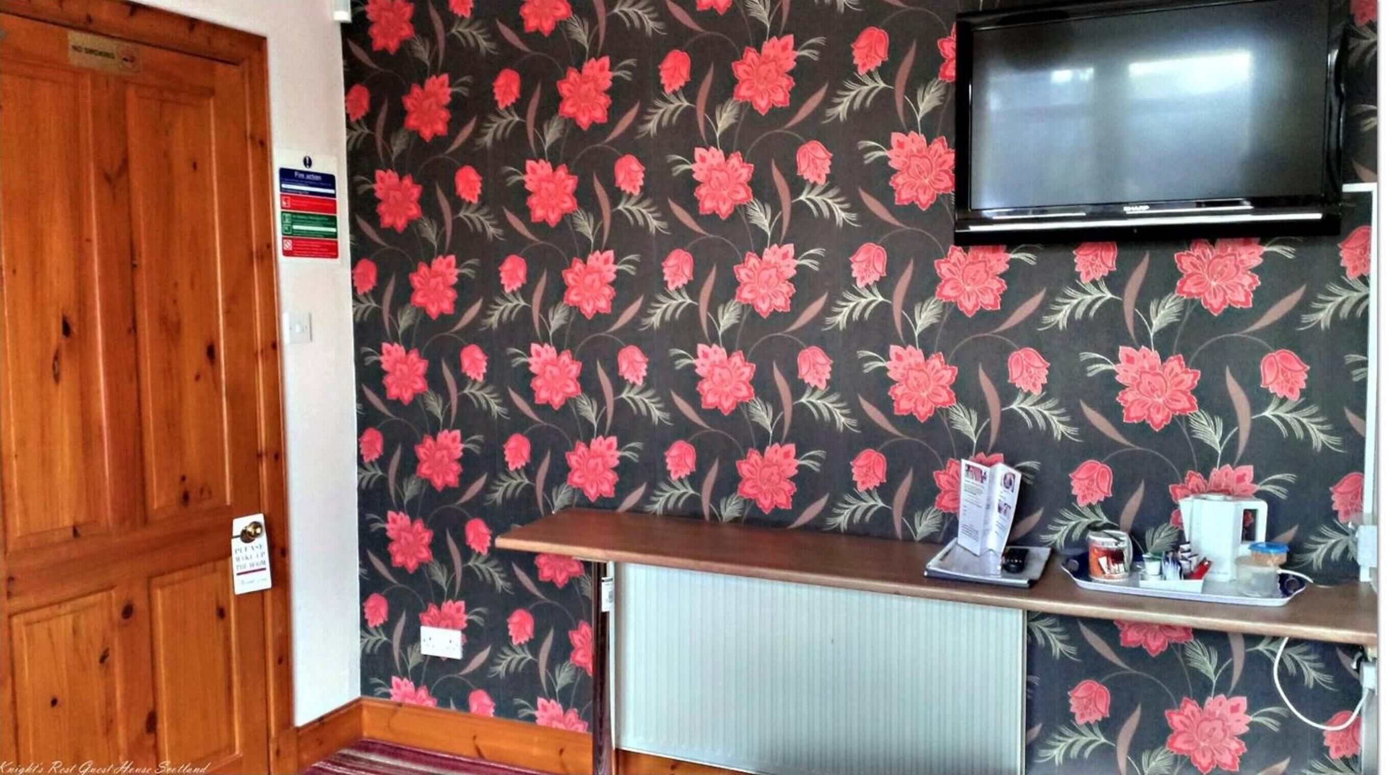 Huoneessa on puinen ovi, seinälle asennettu televisio, pieni pöytä, jossa on vedenkeitin ja kuppeja, sekä patteri.