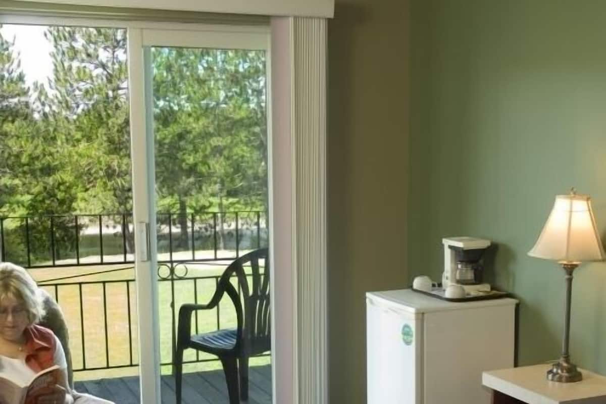 A room with a bed, a chair, a table with a lamp, a refrigerator, and a sliding glass door to an outdoor area.
