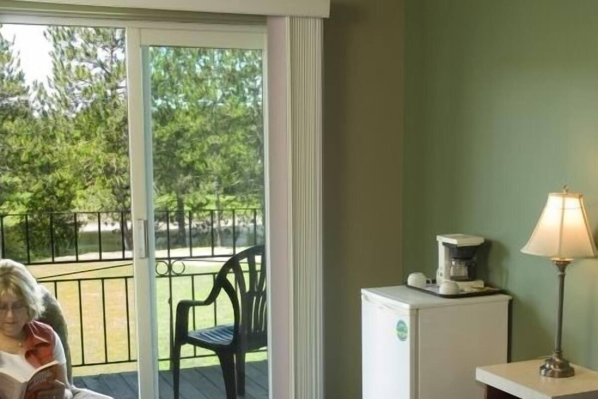 A room with a bed, a chair, a table with a lamp, a refrigerator, and a sliding glass door to an outdoor area.