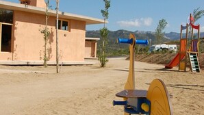 Ein Spielplatz mit Rutsche und Klettergerüst, ein Gebäude mit sandfarbener Fassade und eine gebirgige Landschaft im Hintergrund.