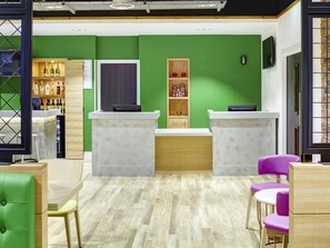 A reception area with a green wall, two reception desks, a wooden cabinet with bottles, and purple chairs. - Ibis Styles Glasgow Centre George Square (Glasgow)