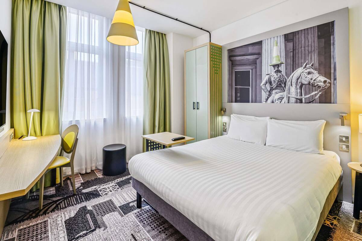 A hotel room with a bed, a desk, a chair, a lamp, a trash bin, and a large painting on the wall.