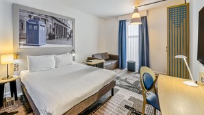 A hotel room with a large bed, a desk with a chair, a sofa, a TV, and a painting of a blue telephone booth. - Ibis Styles Glasgow Centre George Square (Glasgow)