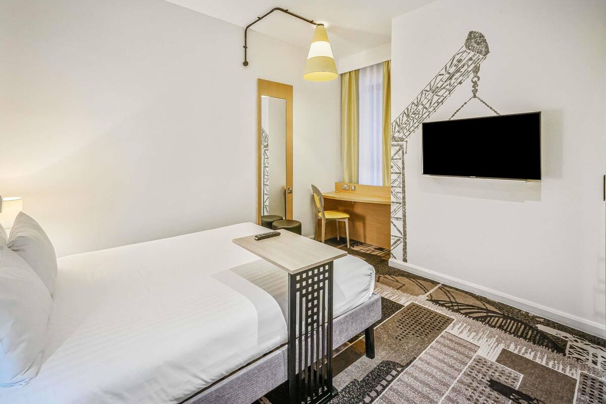 A hotel room with a bed, a desk with a chair, a TV, and a patterned rug.