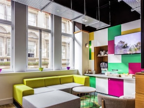 A modern interior with large windows, a sofa, a small table, and a kitchen area. - Ibis Styles Glasgow Centre George Square (Glasgow)