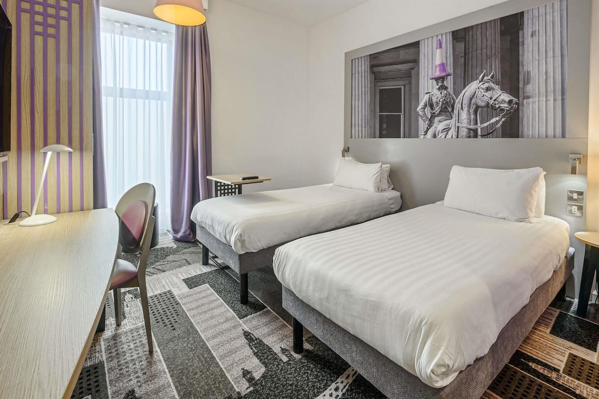 A hotel room with two beds, a desk, a chair, a lamp, and a large window with curtains.