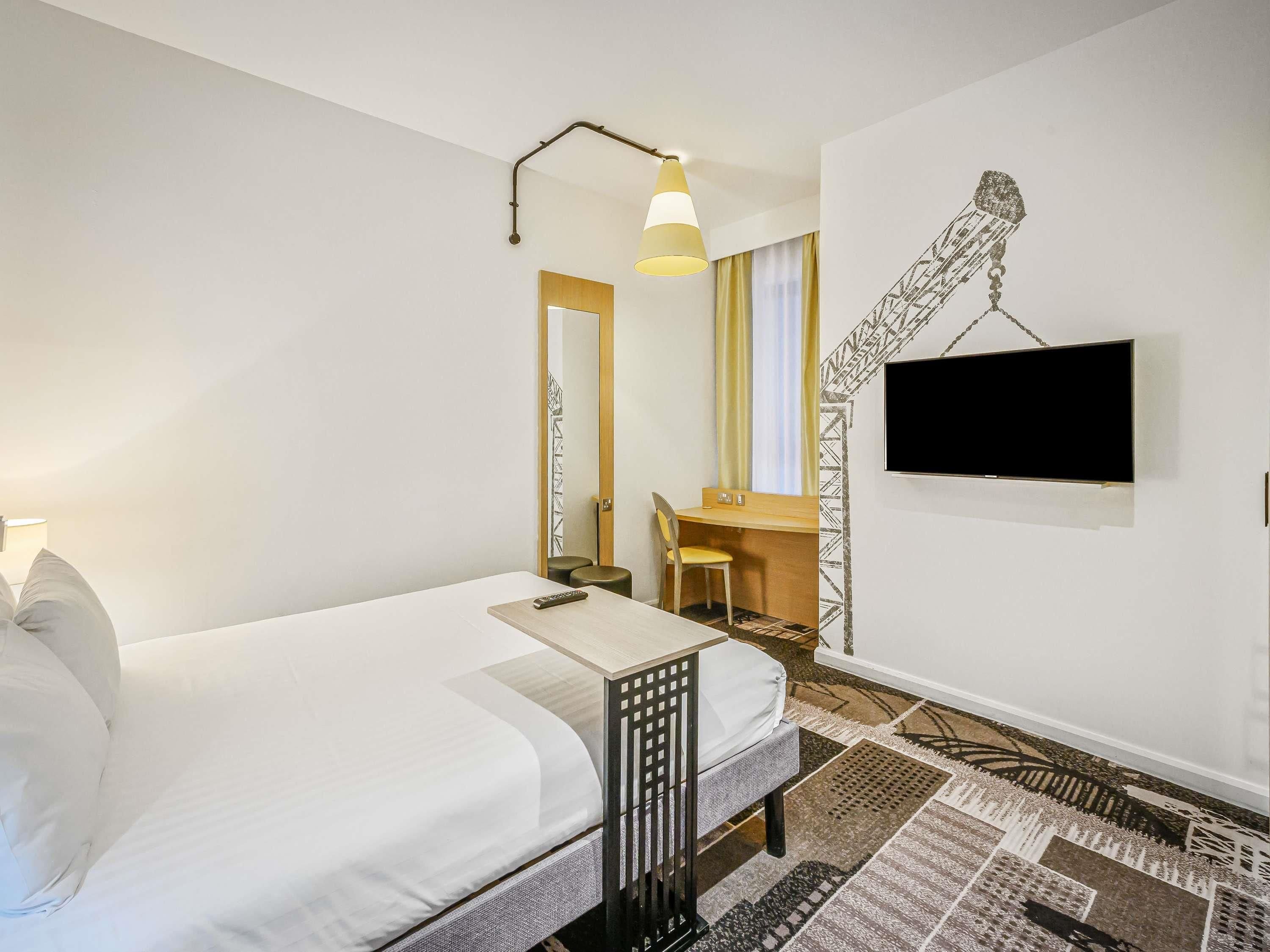 A hotel room with a bed, a desk with a chair, a TV, and a patterned rug.