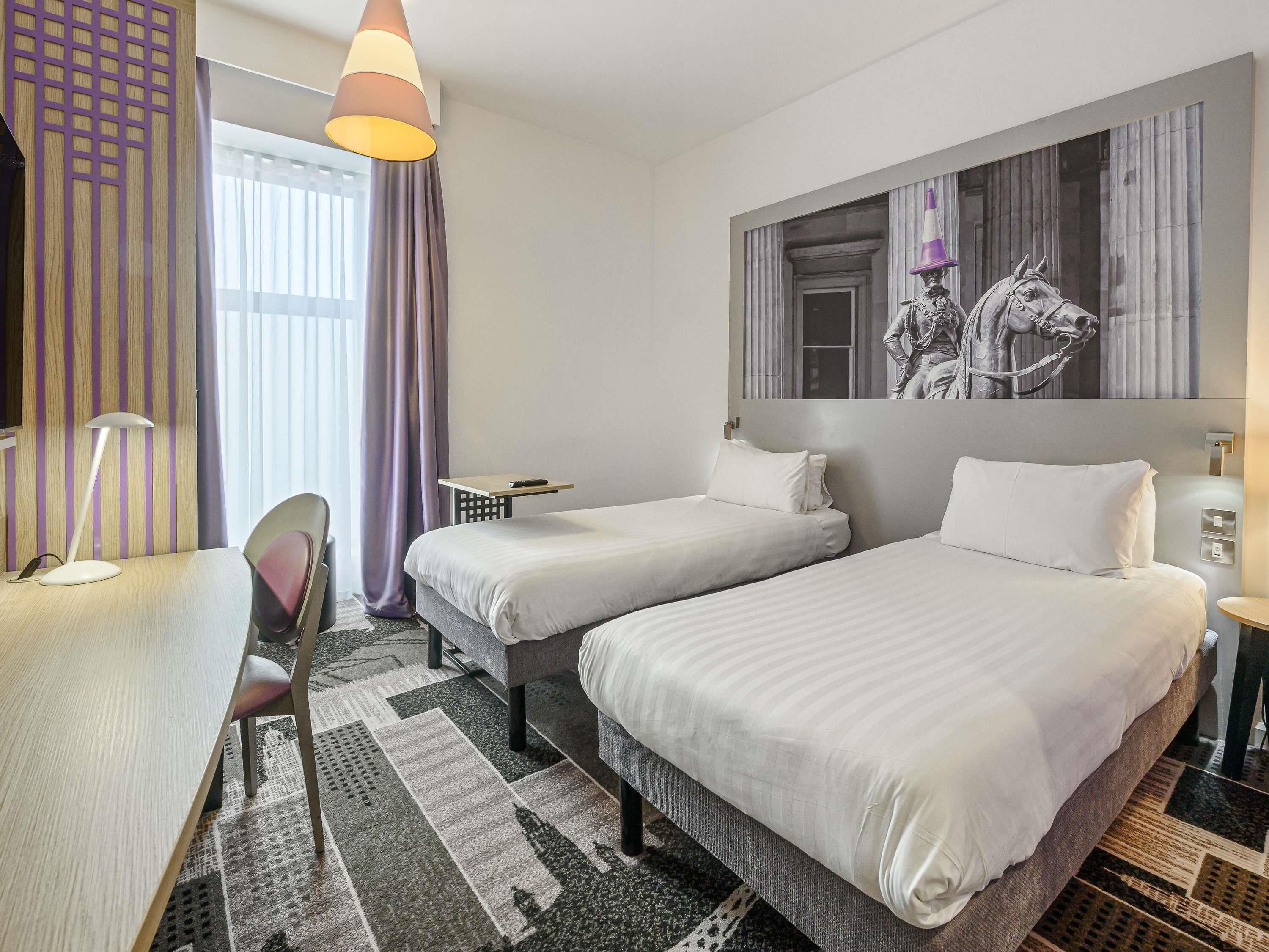 Une chambre d’hôtel avec deux lits, un bureau, une chaise, une lampe et une grande fenêtre avec des rideaux.