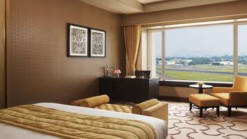 A hotel room with a large bed, a desk with a lamp, a chair, and a view of the airport.