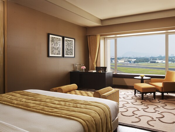 A hotel room with a large bed, a desk with a lamp, a chair, and a view of the airport. - Taj Santacruz (Mumbai)