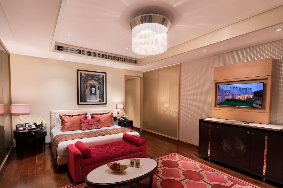 Una habitación de hotel con una cama grande, un sofá rojo, una mesita redonda con fruta, un televisor y una araña.