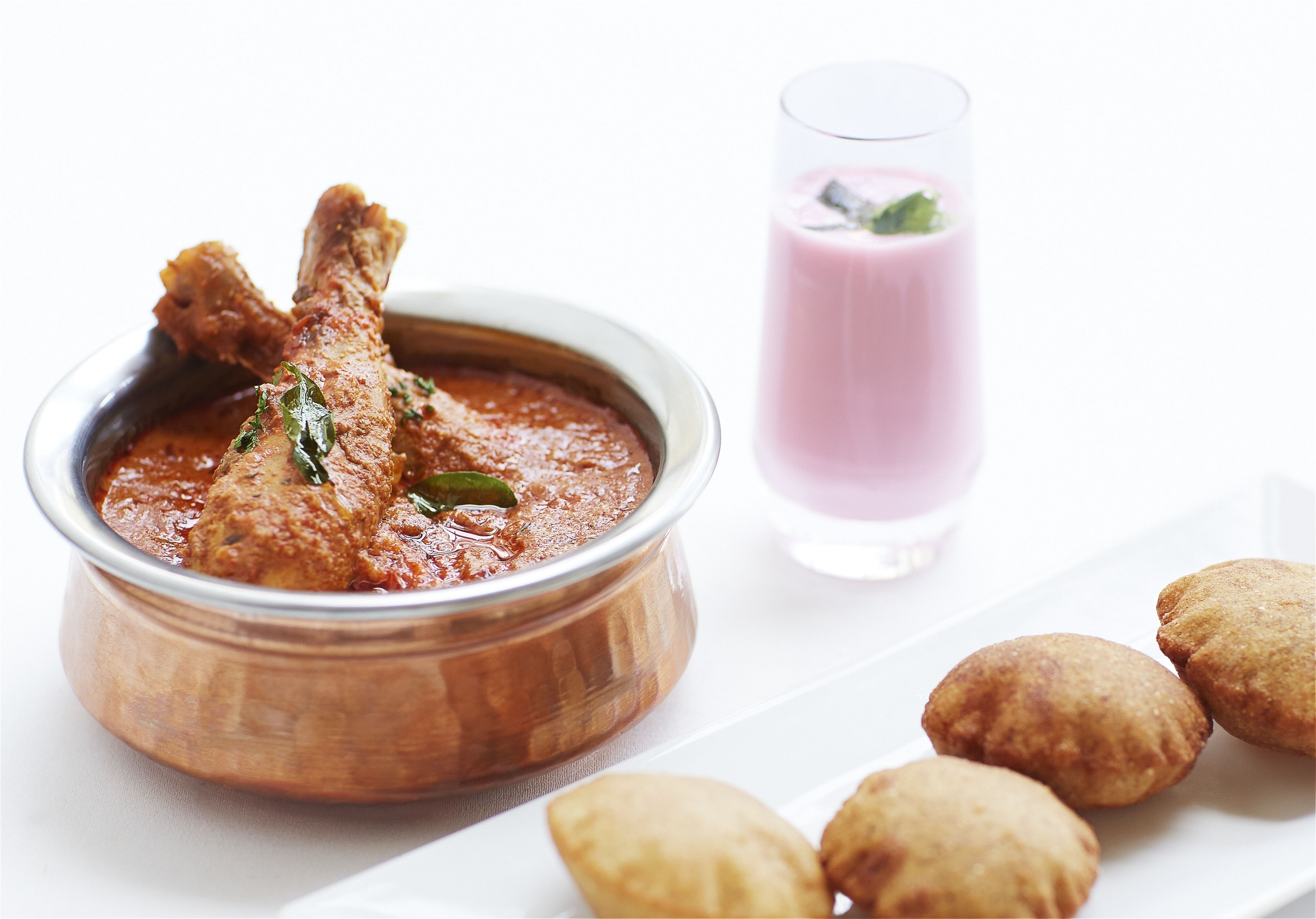 Un tazón de curry de pollo con un vaso de bebida rosada y aperitivos fritos sobre un plato blanco.