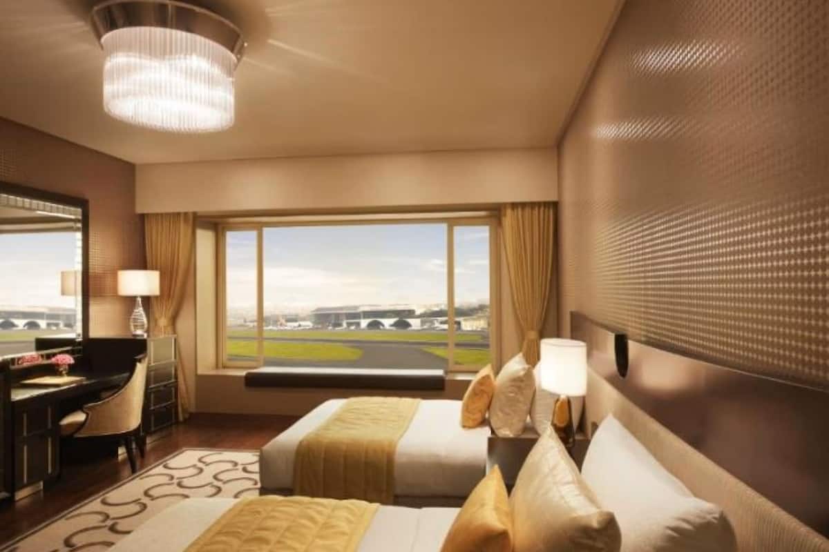 Habitación de hotel con dos camas, un escritorio, una silla, una lámpara y un ventanal con vista al aeropuerto.