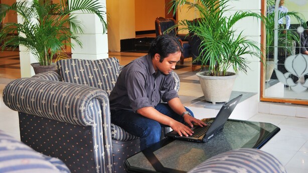 A man sitting on a patterned sofa using a laptop in a lobby with potted plants and a glass wall. - Sepinggan Hotel (Balikpapan)