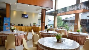 A hotel dining area with round tables covered with patterned tablecloths, beige chairs, and potted plants. A television is mounted on the wall in the background. - Sepinggan Hotel (Balikpapan)