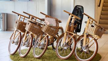 Una exhibición de bicicletas antiguas con cestas de mimbre, sobre una alfombra verde en una habitación con una escalera y un cubo de basura al fondo.