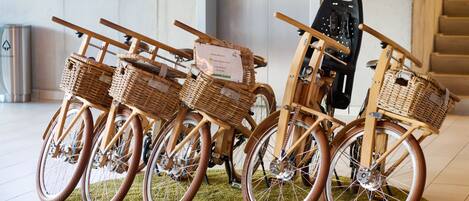 Una exhibición de bicicletas antiguas con cestas de mimbre, sobre una alfombra verde en una habitación con una escalera y un cubo de basura al fondo.