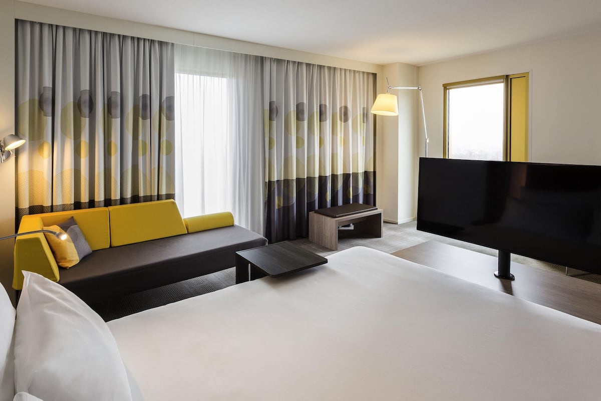 Habitación de hotel con una cama grande, un sofá, una mesita, un televisor y cortinas con un diseño geométrico.